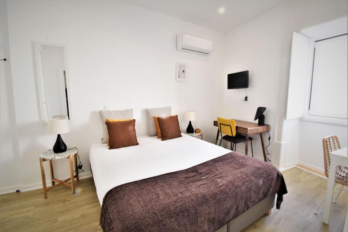 Quarto de hotel moderno com uma cama grande, uma escrivaninha, uma cadeira, uma televisão e um aparelho de ar condicionado.