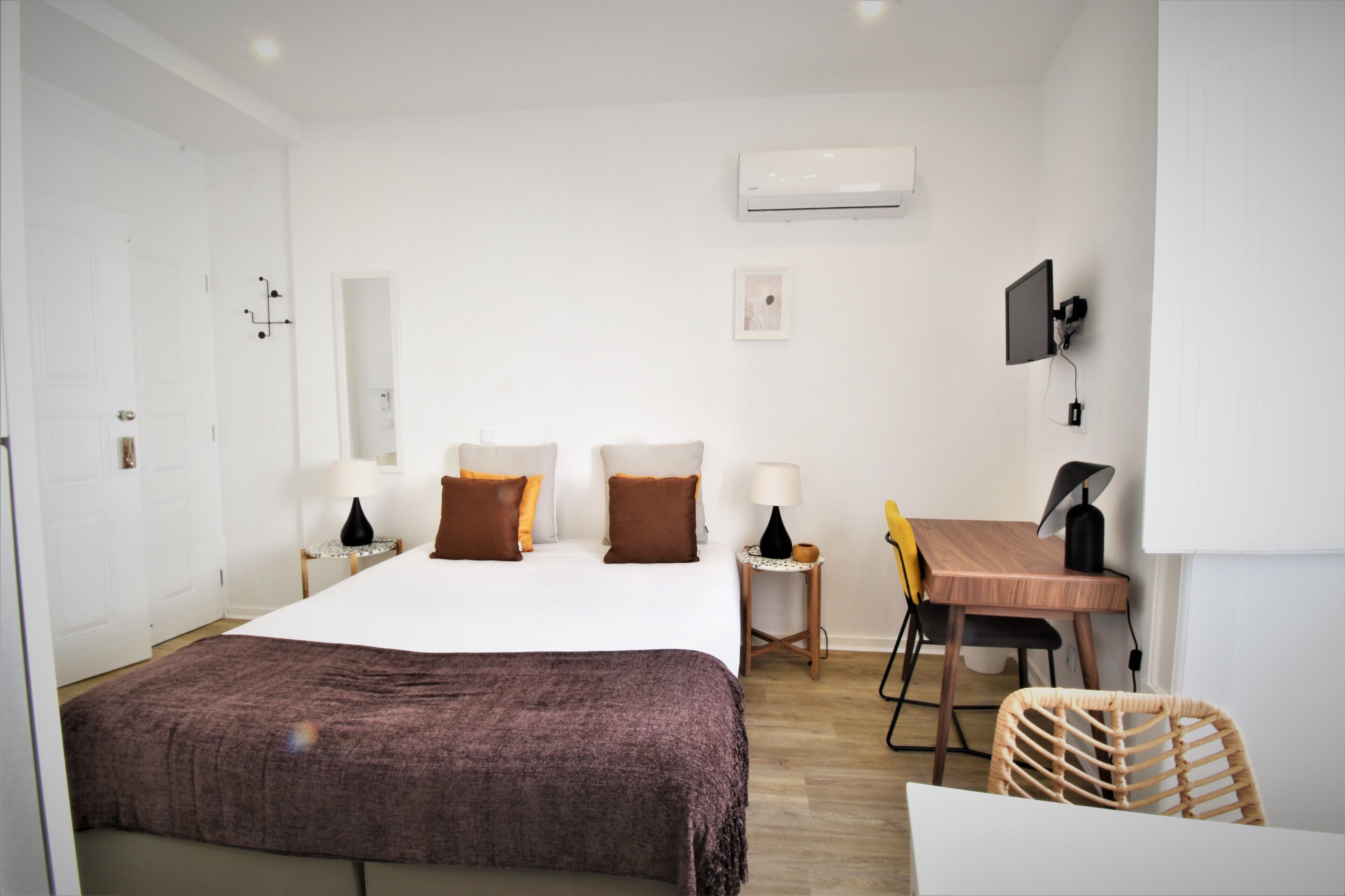 A hotel room with a bed, bedside tables, a desk, a chair, and an air conditioning unit on the wall.