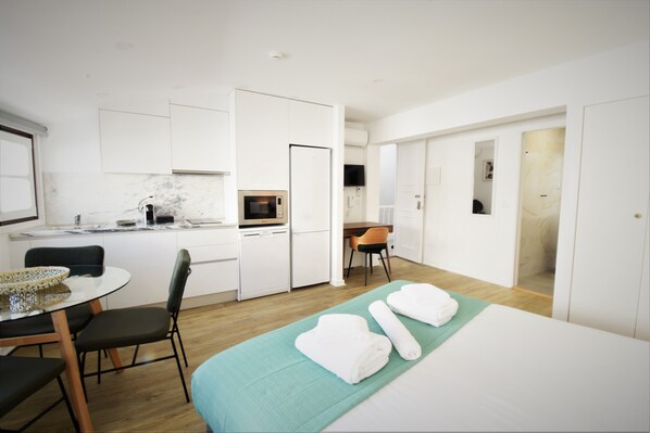 A modern hotel room with a bed, a dining table with chairs, a kitchen area with a microwave, and a TV.