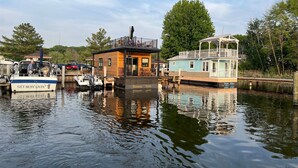 Une propriété située au bord de l’eau, avec un bateau amarré, un petit bâtiment en bois et une plus grande structure bleue dotée d’un balcon.