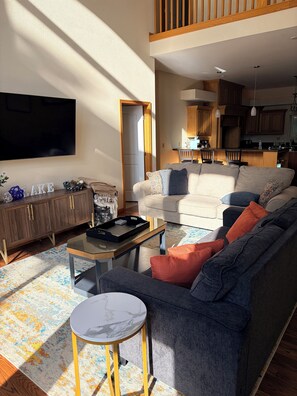 A living room with a sofa, coffee table, and TV. There is a kitchen in the background.