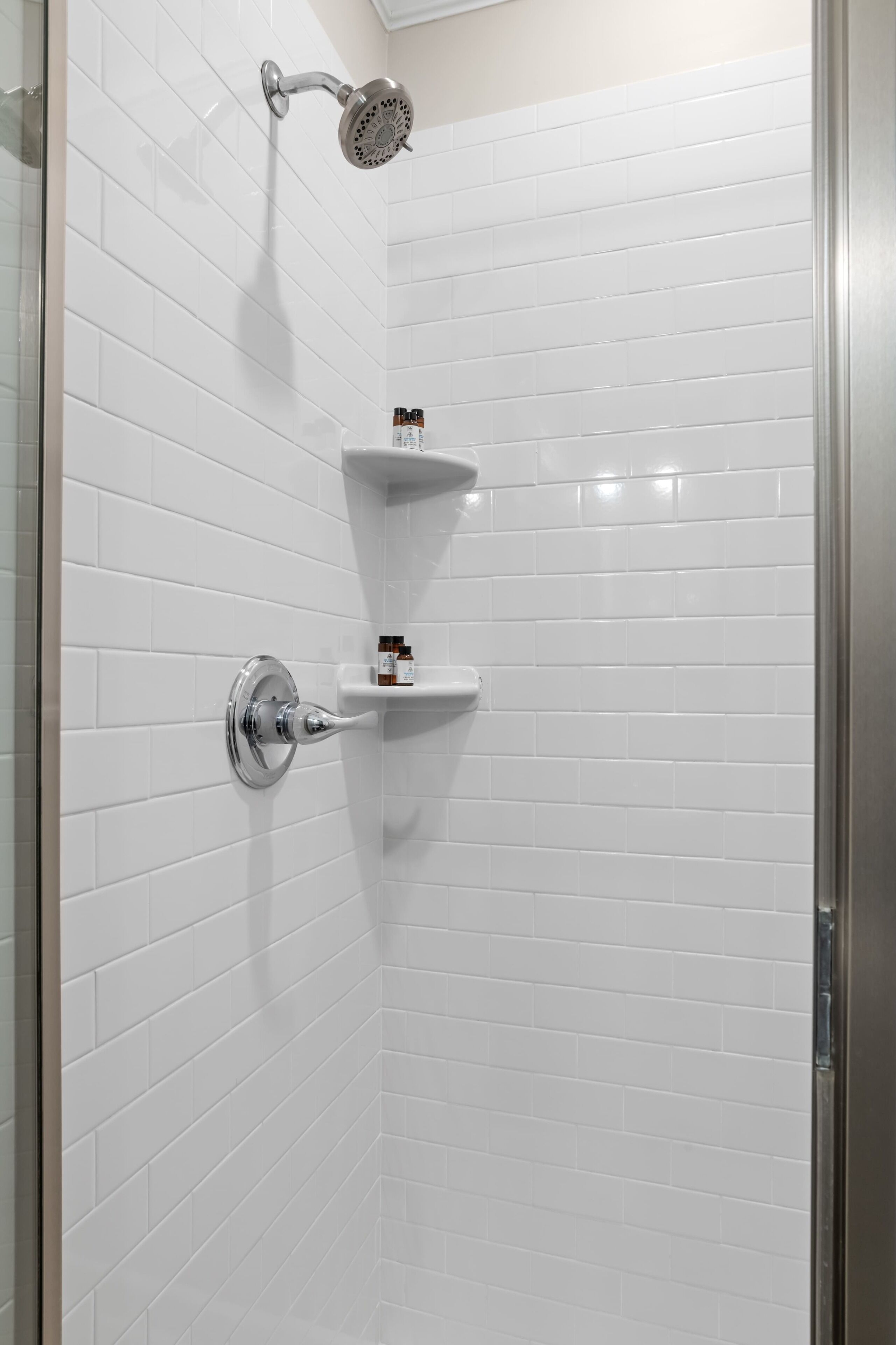 A shower with white tiles, a handheld showerhead, and two small shelves with toiletries.
