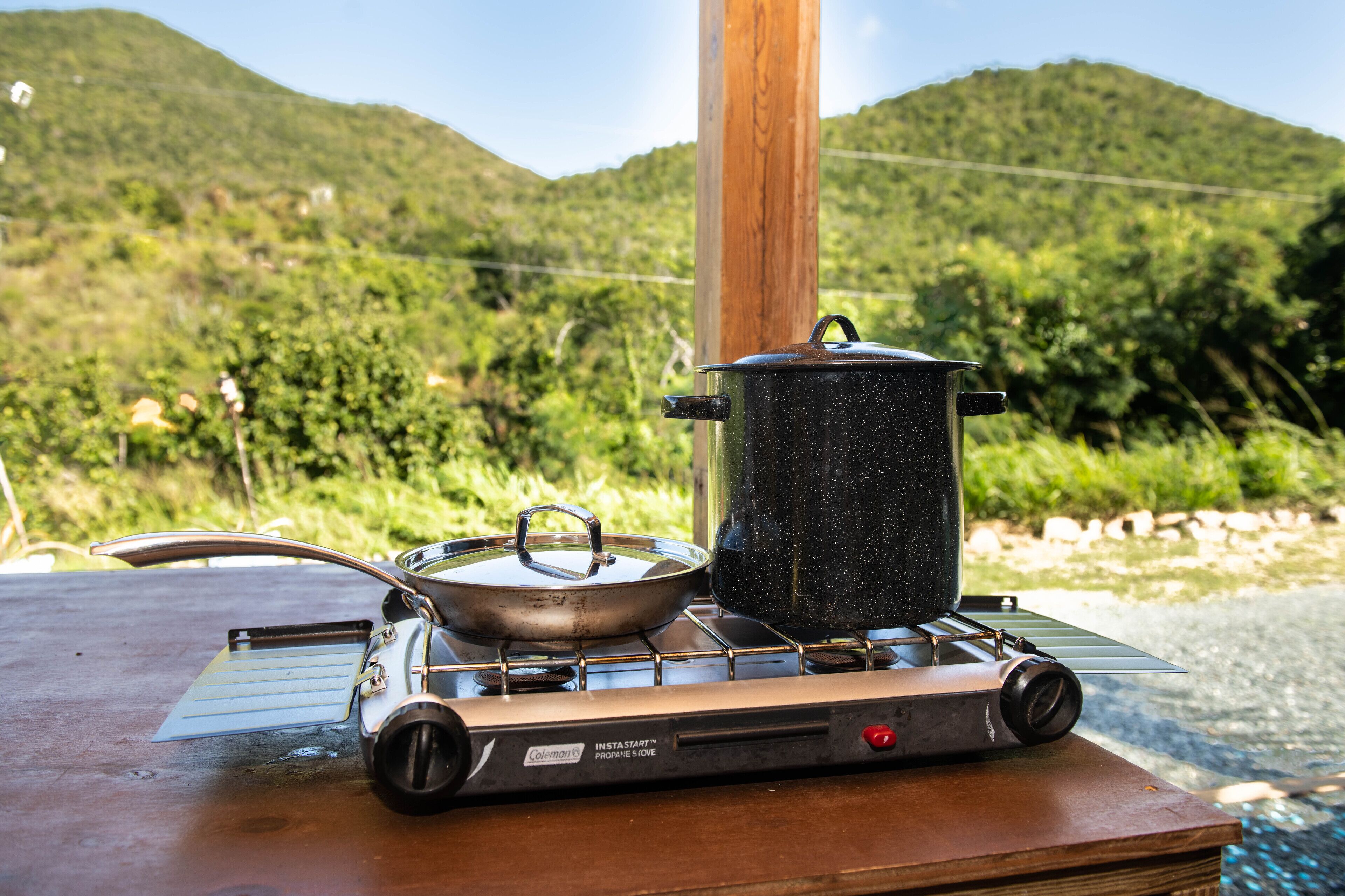 Un réchaud Coleman portable, avec une casserole et une poêle posées dessus, est installé sur une table en bois à l’extérieur, avec une chaîne de montagnes en arrière-plan.