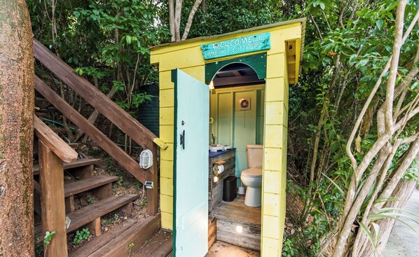 A small, yellow wooden structure labeled "Welcome to Lameshlu Cottage" with a wooden staircase leading up to it. Inside, there is a toilet and a small sink.
