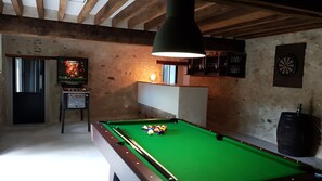 A pool table in a room with wooden beams on the ceiling, a dartboard, and a television displaying a game.