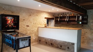 Uma área de bar com uma máquina de fliperama antiga, uma estante de madeira com garrafas e um balcão branco.