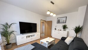 Una sala de estar moderna con un televisor de pantalla plana grande, un sofá gris oscuro, una mesa de centro blanca y un mueble blanco minimalista.