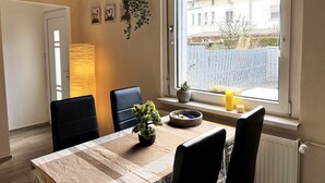 Un comedor con una mesa, sillas, una planta colgante, un ventanal con vistas a edificios y una puerta.