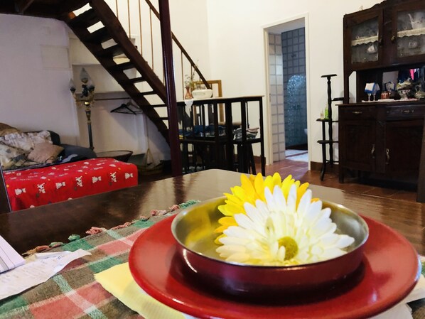 Une table à manger recouverte d’une nappe rouge, un vase contenant une fleur blanche et un escalier en arrière-plan.