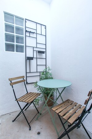 Un pequeño espacio al aire libre con una mesa y dos sillas, un ventanal con vidrio esmerilado y una planta en maceta.