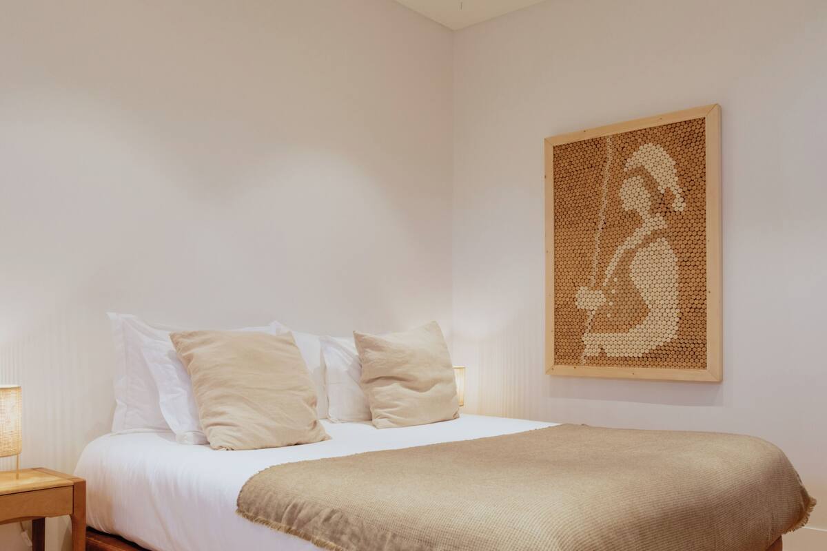 A bedroom with a bed, pillows, a blanket, a bedside table, a lamp, and a wall art piece.