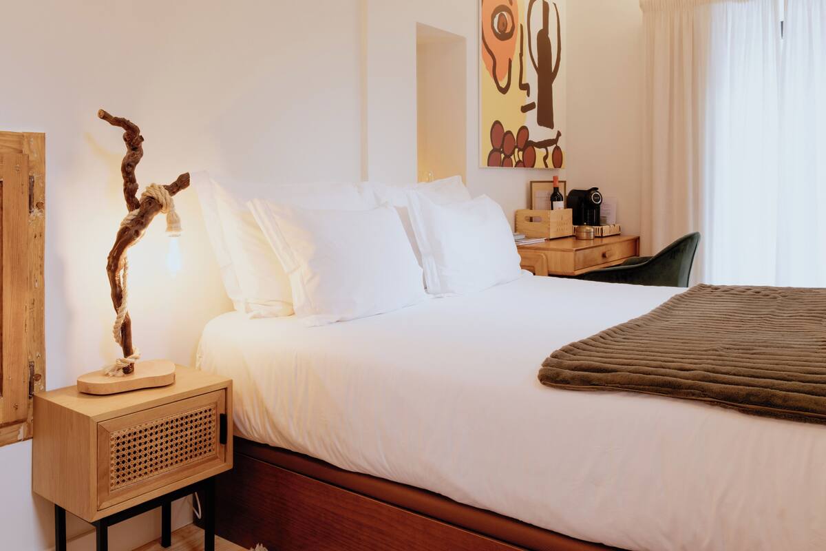 A neatly made bed with white linens, a wooden bedside table with a lamp, a decorative branch, and a painting on the wall.