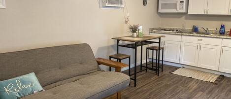 A compact living space with a sofa, a small table, a kitchen area with white cabinets, and an air conditioning unit.