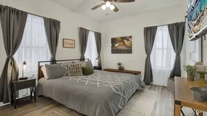 Une chambre à coucher comprenant un lit, un ventilateur de plafond, une table de chevet et une commode, ainsi qu’une fenêtre avec des rideaux.