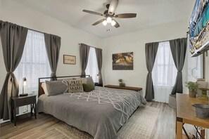 Un dormitorio con cama, ventilador de techo, mesita de noche y ventana con cortinas.