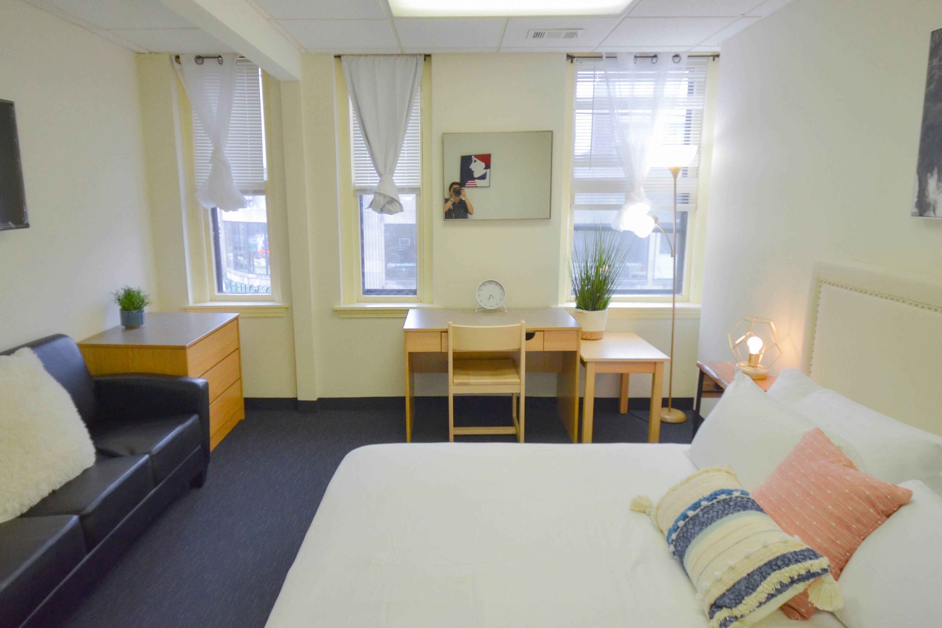 A hotel room with a bed, a desk, a chair, a small table, a nightstand, a lamp, and a window with curtains.