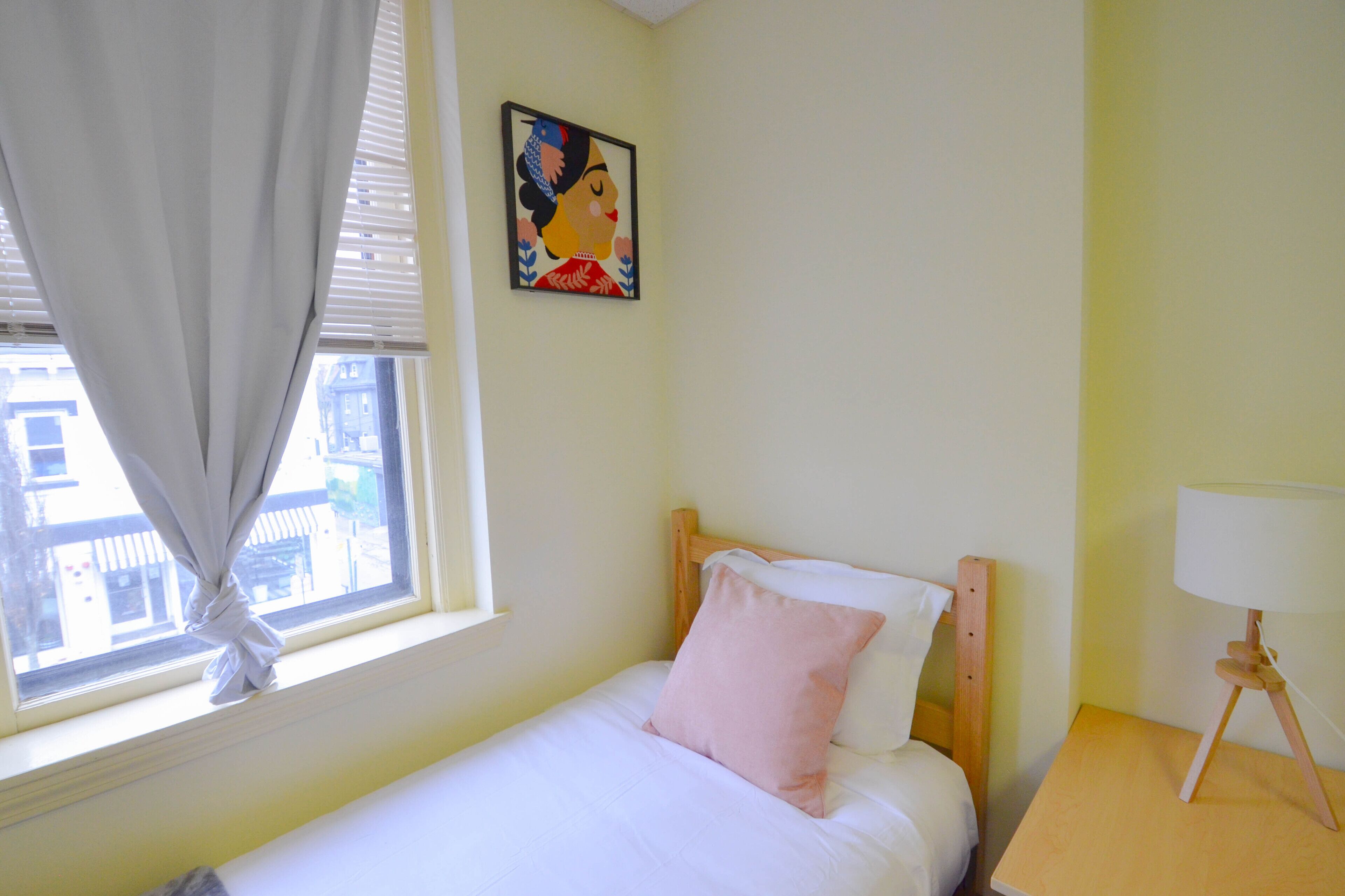 A bedroom with a single bed, a nightstand with a lamp, a window with curtains, and a painting on the wall.