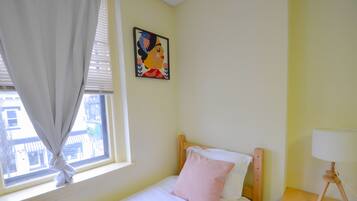 A bedroom with a single bed, a nightstand with a lamp, a window with curtains, and a painting on the wall.