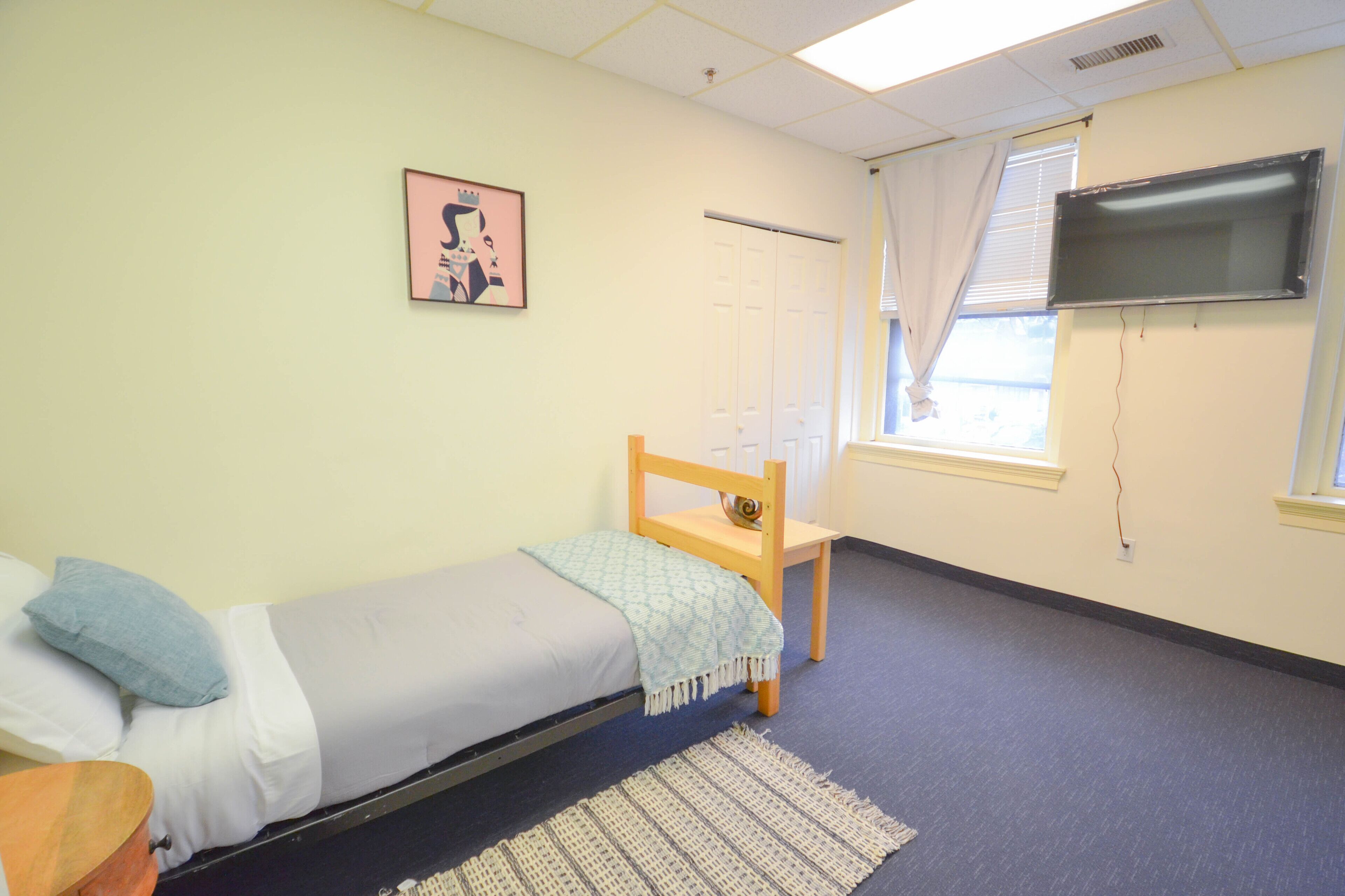 A single-bed room with a bed, a chair, a TV, a window with curtains, and a door.
