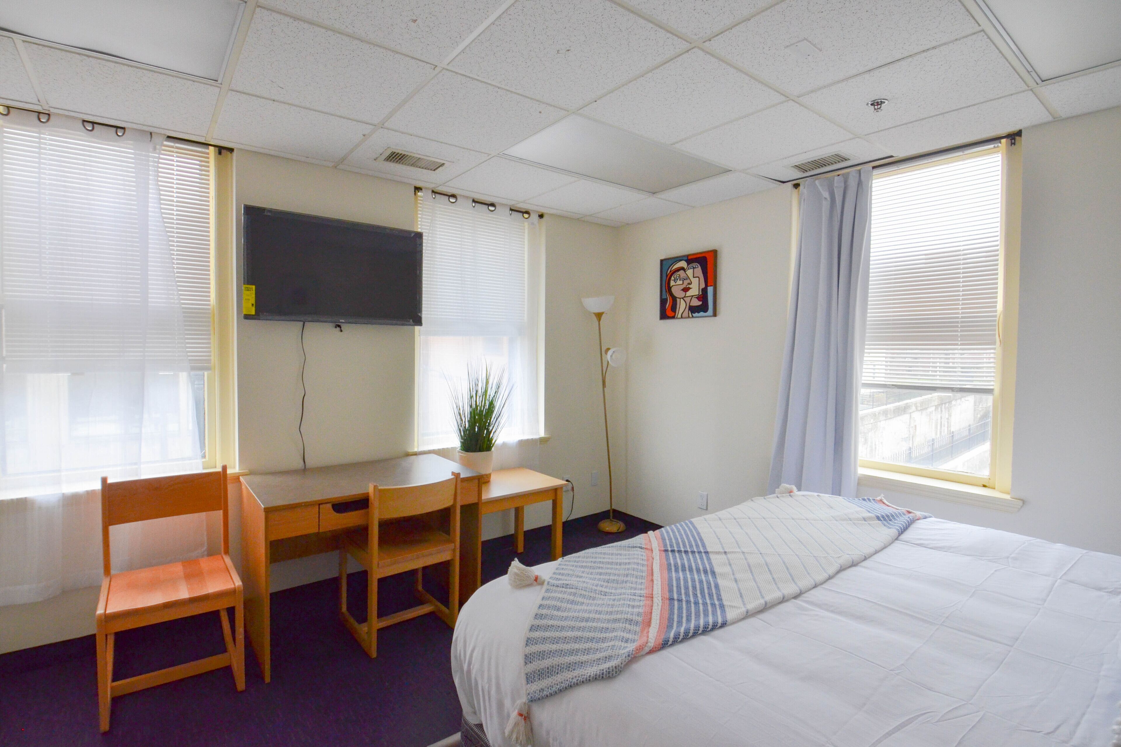 Ein Hotelzimmer mit Bett, Schreibtisch, Stuhl, Fernseher und einem Fenster mit Jalousien.