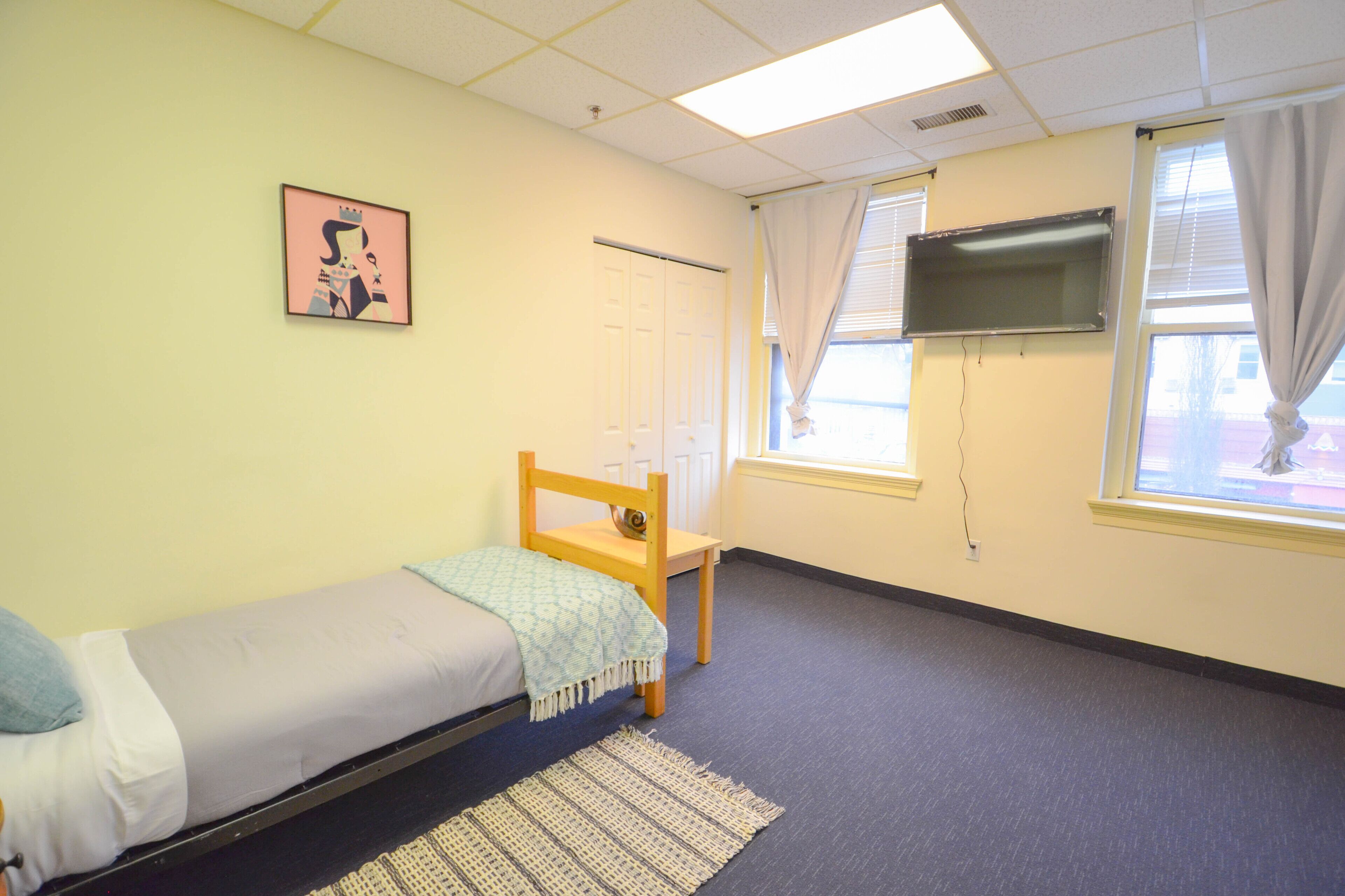 A small, single-bed room with a wooden bed, a small chair, a TV mounted on the wall, and a window with curtains.