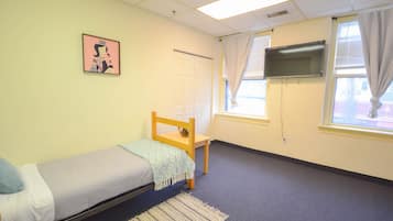 A small, single-bed room with a wooden bed, a small chair, a TV mounted on the wall, and a window with curtains.