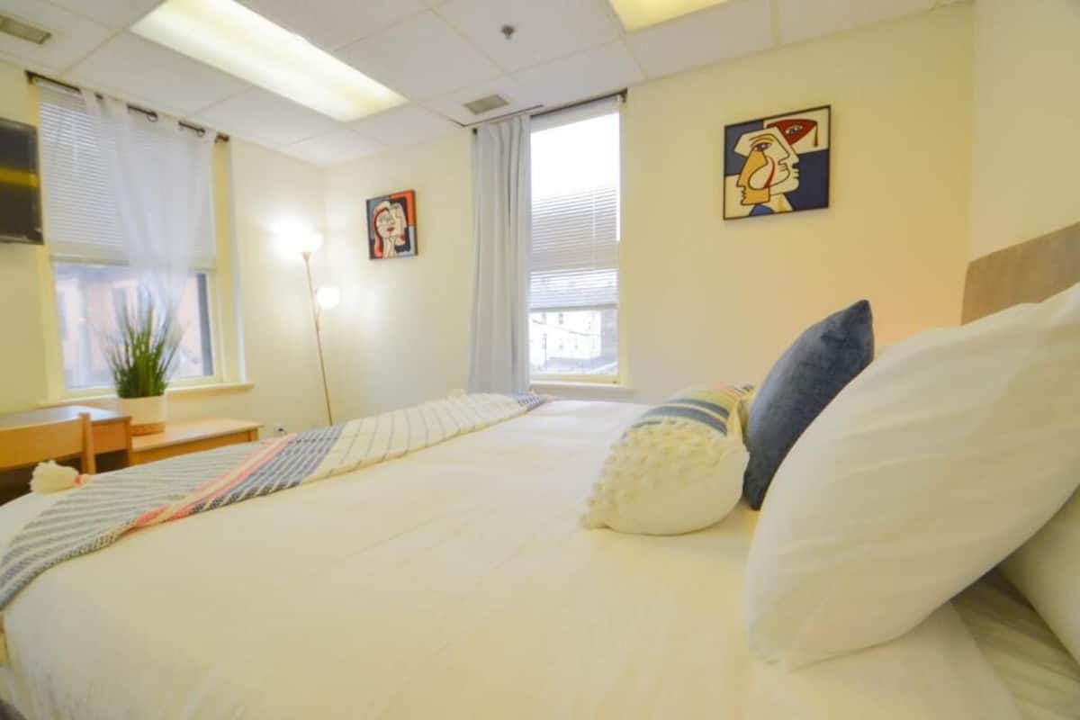 A hotel room with a bed, a desk, a chair, a potted plant, and two paintings on the wall.