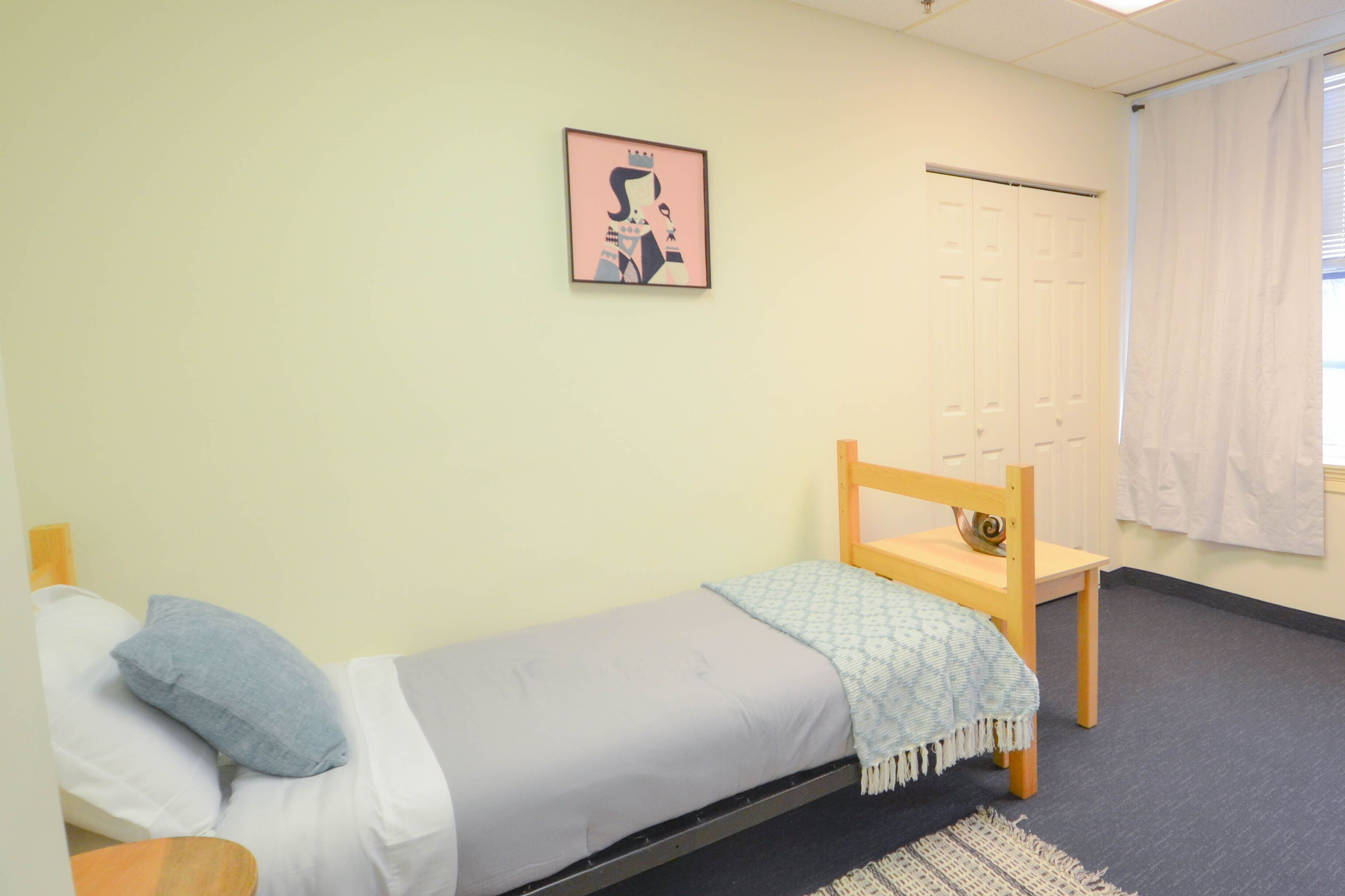 A small, single-bed room with a wooden chair, a framed picture on the wall, and a window with curtains.