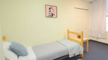 A small, single-bed room with a wooden chair, a framed picture on the wall, and a window with curtains.