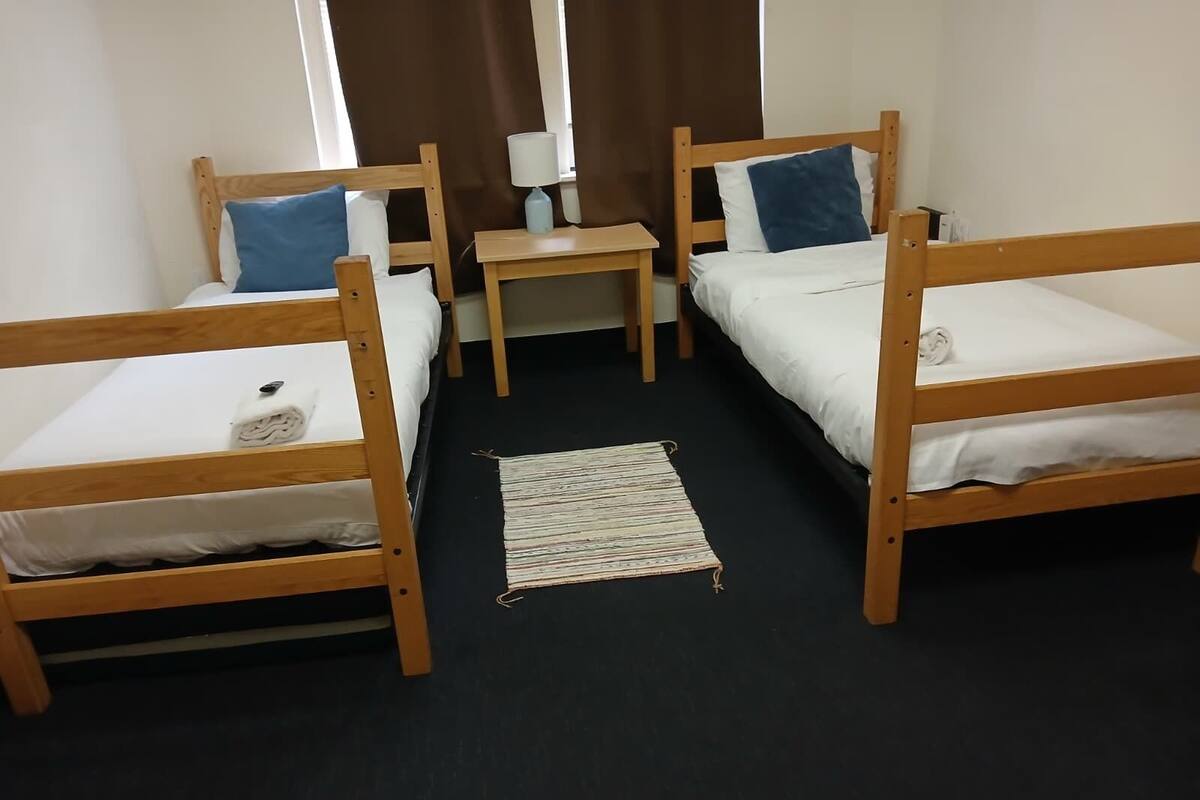 A room with two wooden bunk beds, a small table with a lamp, and a woven rug on the floor.