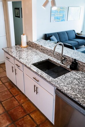 A kitchen with granite countertops, a stainless steel sink, and a dishwasher. There is a blue sofa in the background.