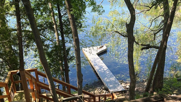 A wooden deck with a wooden staircase leading to a dock over a calm lake, surrounded by trees. - Road America Adjacent Lake House with indoor pool (Plymouth)