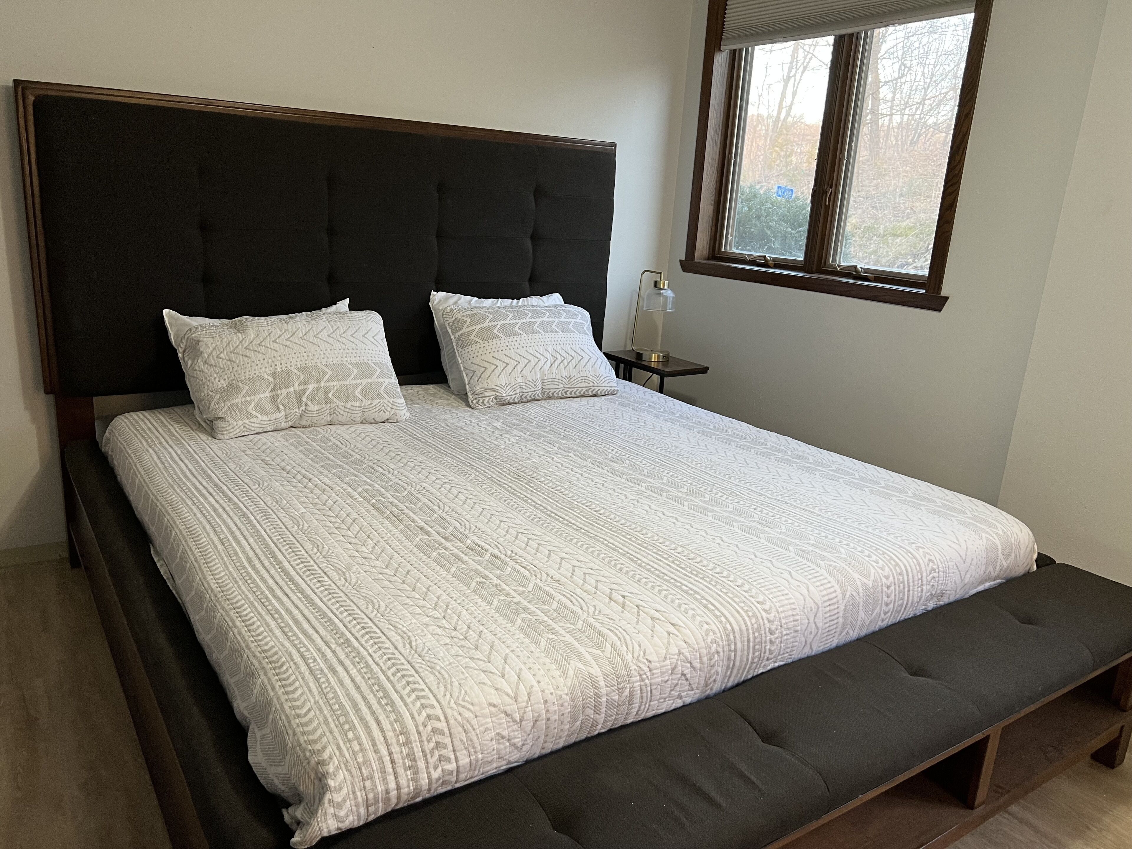 A bed with a dark headboard, two pillows, a bedside table with a lamp, and a window with blinds.