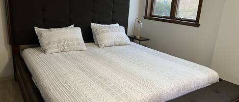 A bed with a dark headboard, two pillows, a bedside table with a lamp, and a window with blinds.