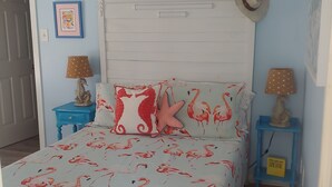 Un dormitorio con una cama que presenta un edredón estampado con flamenco, un cabecero blanco y almohadas a juego. Hay dos mesitas de noche con lámparas, una con una pantalla de lunares y la otra con un sombrero.
