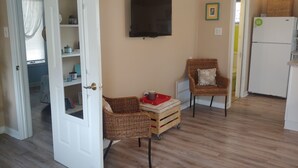 Una habitación con piso de madera, un televisor montado en la pared, un sillón de mimbre con cojín, una pequeña mesa de madera con una bandeja roja y un refrigerador blanco.
