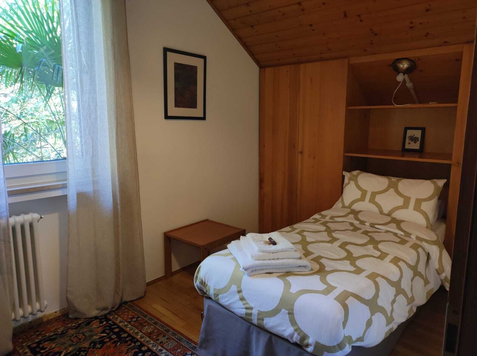 A bedroom with a bed, a small wooden nightstand, a radiator, a window with curtains, and a wooden ceiling.