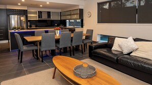 Een moderne keuken met een centraal eiland, een eethoek met stoelen, een houten salontafel en een zwarte leren bank met een wit kussen.