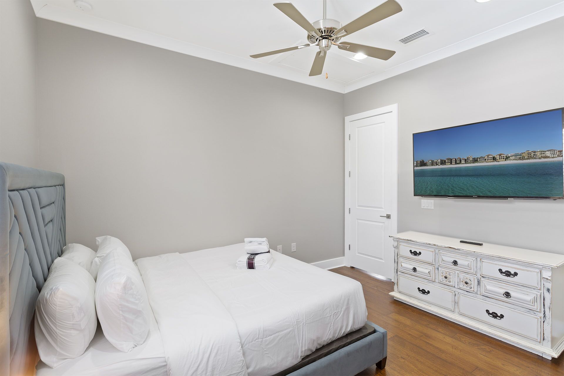 A bedroom with a bed, a ceiling fan, a TV on the wall, and a dresser.