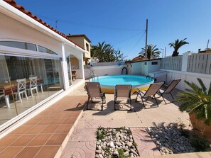 A swimming pool with lounge chairs, a glass-enclosed dining area, and a tiled patio.