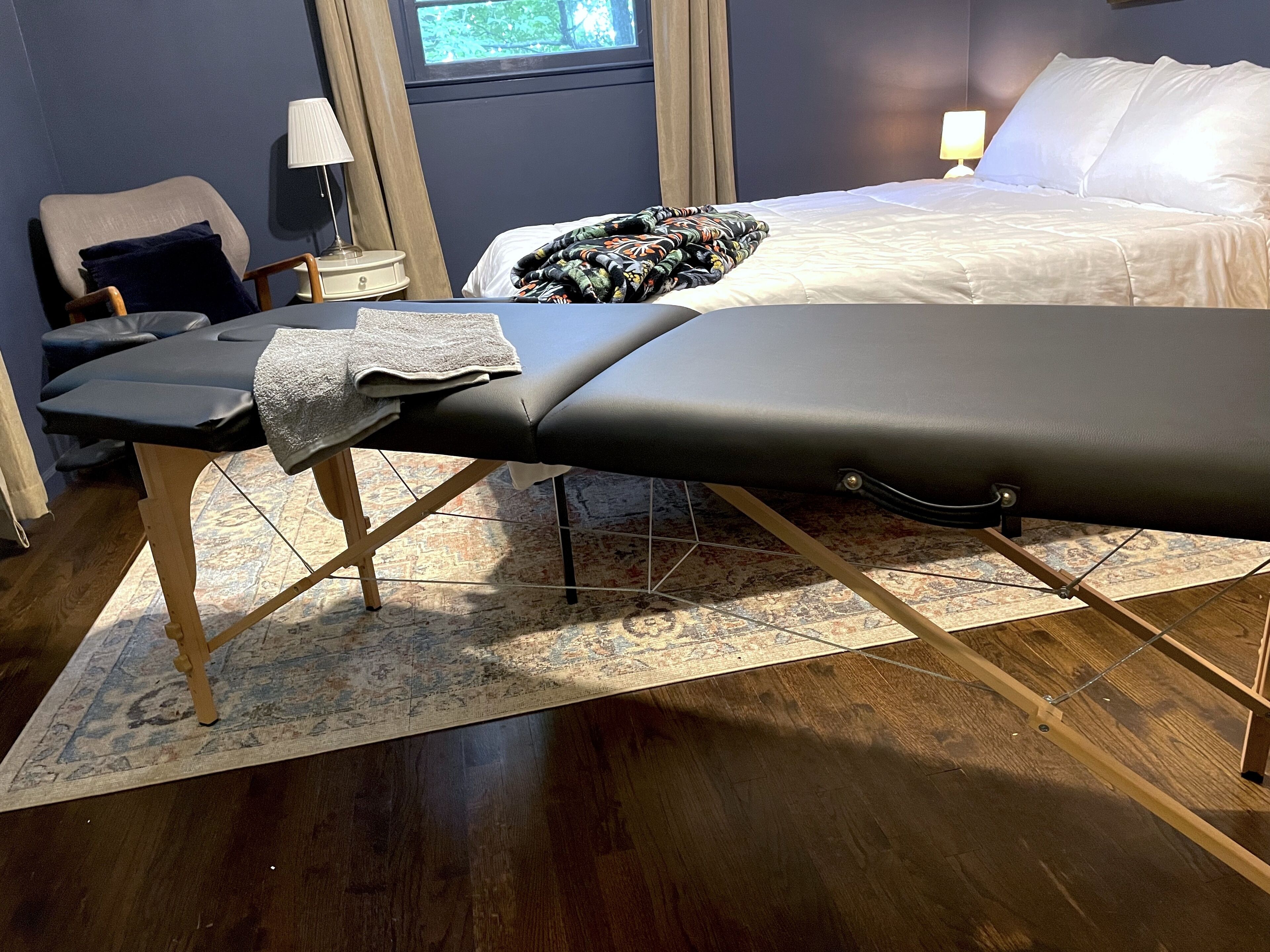 Una mesa de masaje en una habitación con una cama, una silla, una lámpara y una ventana.