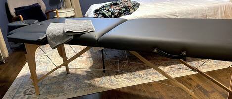 Una mesa de masaje en una habitación con una cama, una silla, una lámpara y una ventana.
