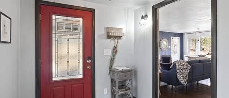 Una puerta roja con un panel de vidrio, un letrero de bienvenida, una pequeña mesa con un espejo y un sofá azul en la habitación contigua.
