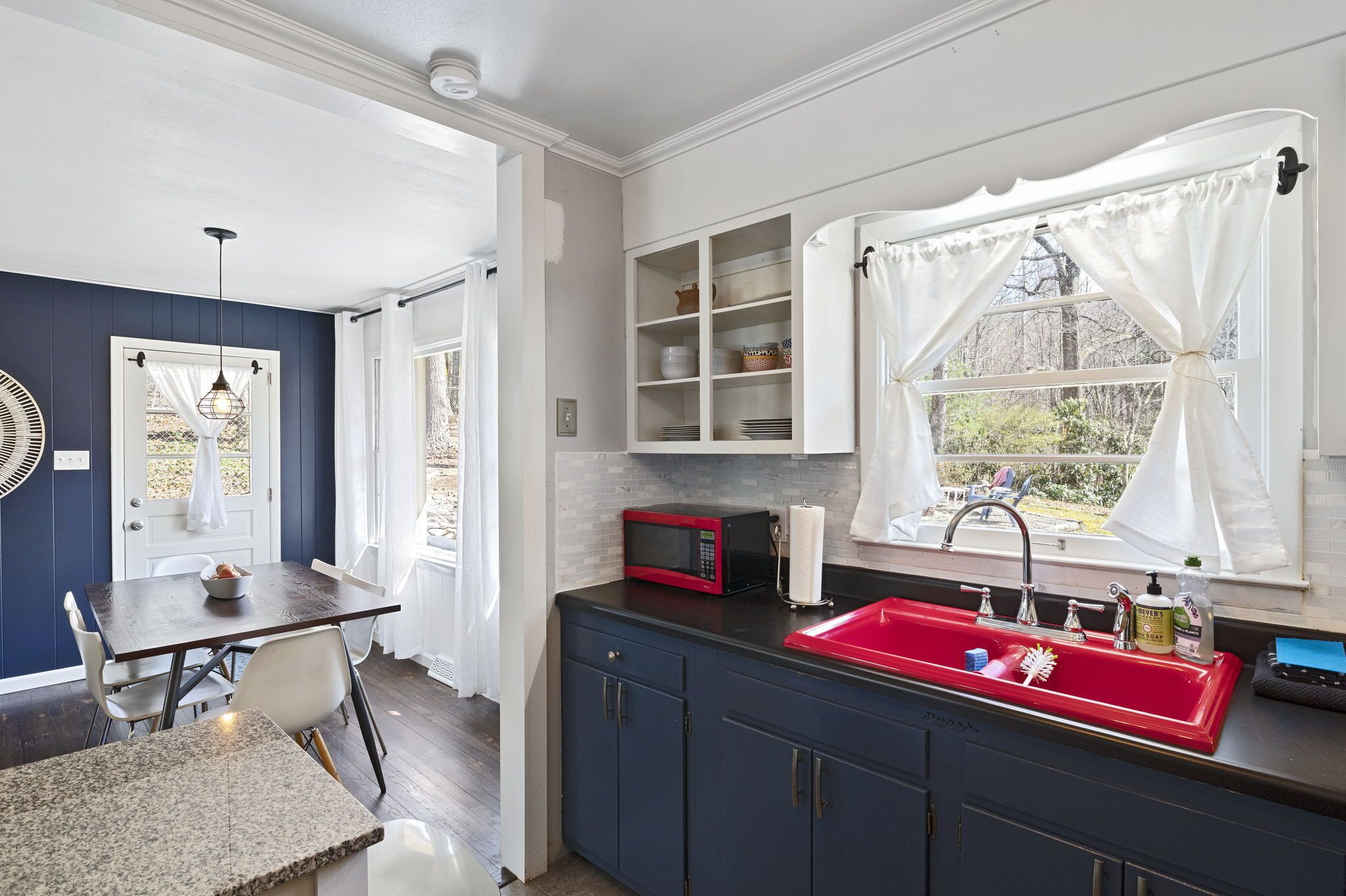 A kitchen with a red sink, white cabinets, and a microwave. There is a window with white curtains and a view of greenery outside.