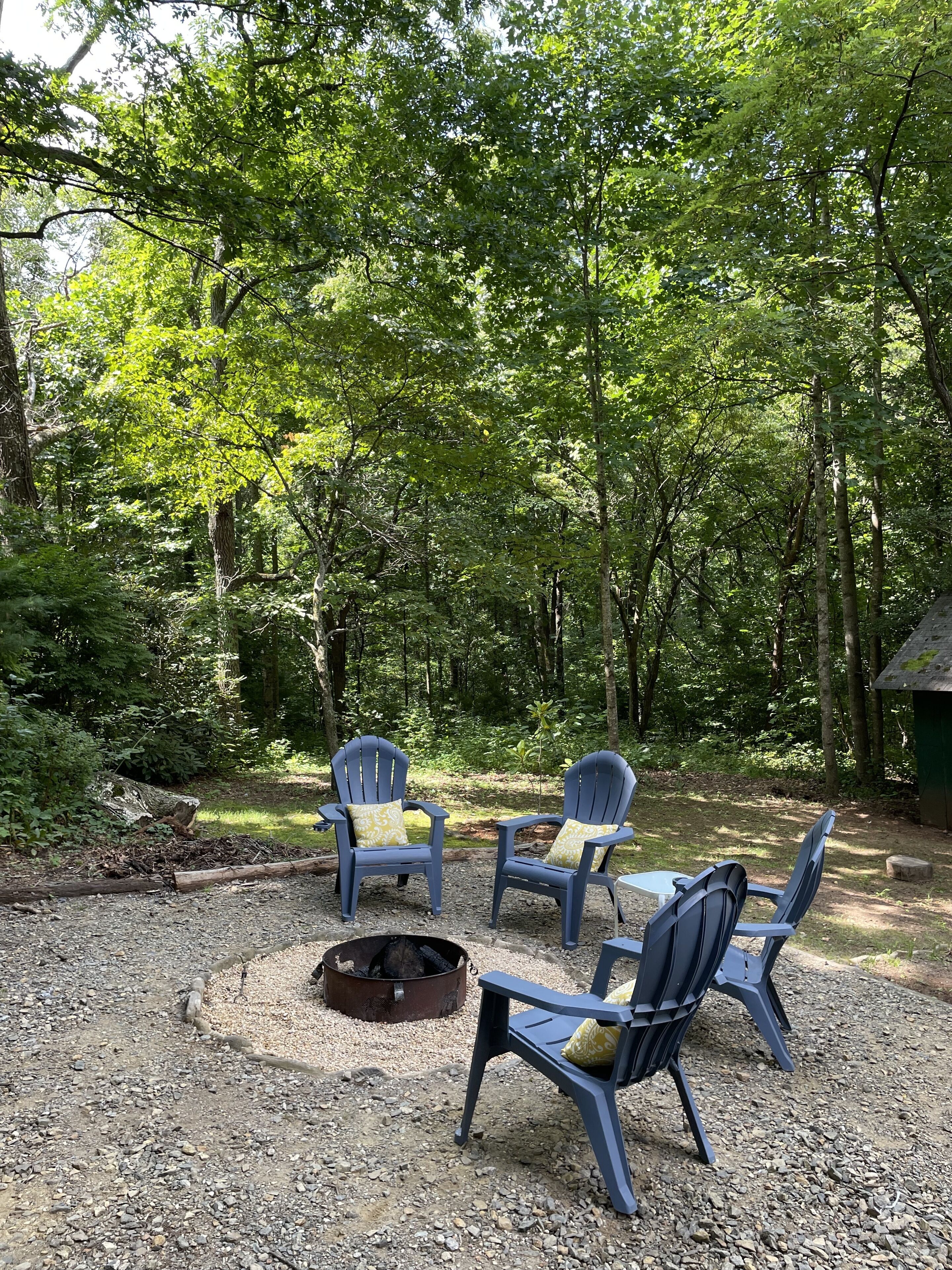 A forested outdoor area with four Adirondack chairs and a fire pit.