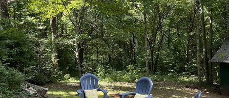 A forested outdoor area with four Adirondack chairs and a fire pit.