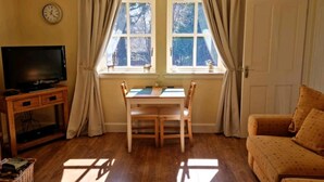Ein Wohnzimmer mit Fernseher auf einem Holzgestell, einem Esstisch mit Stühlen, einer Couch und einem Fenster mit Vorhängen.