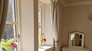 Ein Zimmer mit Fenster, einem Schminktisch mit Spiegel und zwei Vasen mit Blumen auf dem Fensterbrett.