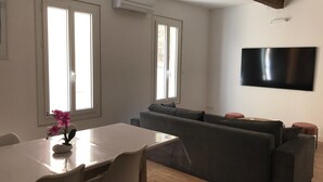 Ein modernes Wohnzimmer mit Essbereich, großen Fenstern, einem wandmontierten Flachbildfernseher und einem Holzbalken an der Decke.