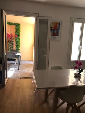 Un espace repas comprenant une table et des chaises, une cloison en verre et une vue sur un balcon agrémenté de plantes et de fleurs.